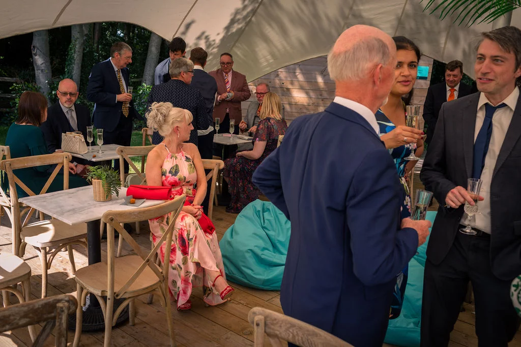 Guests enjoy champagne and conversation inside a bright marquee reception featuring wooden flooring and relaxed seating.