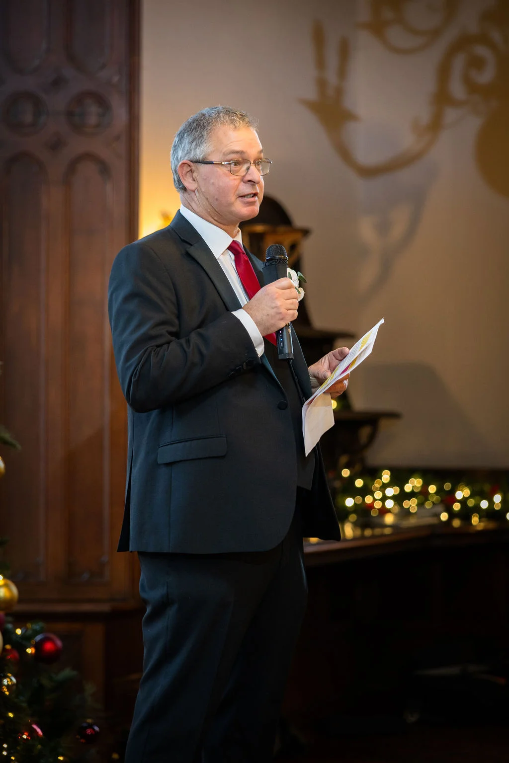 An older man in a dark suit and red tie delivers a wedding speech holding a microphone and notes.