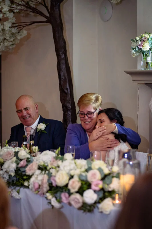 An emotional mother of the bride embraces the bride warmly at the top table during the wedding reception speeches, with floral arrangements in the foreground.
