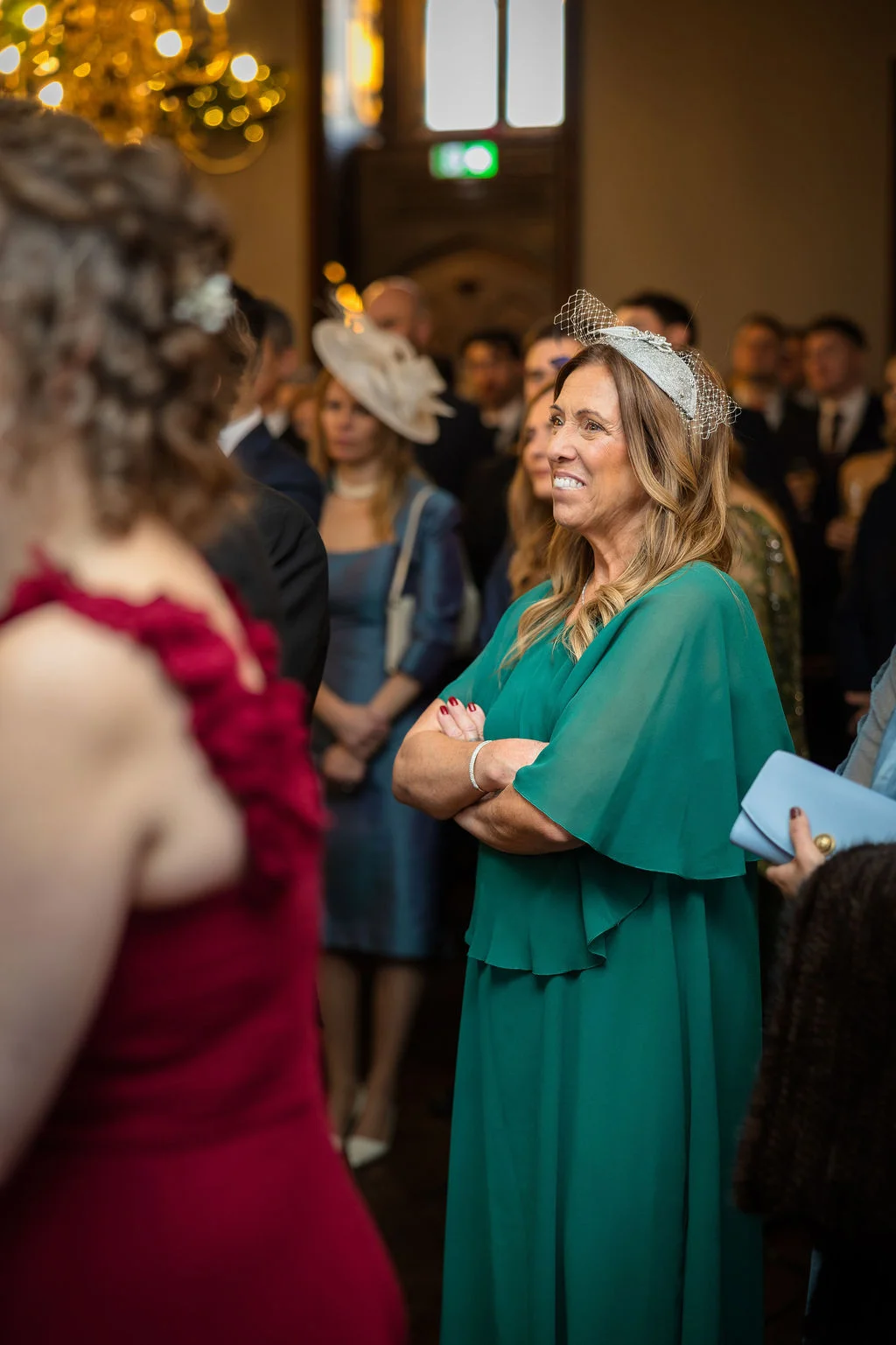 An elegant female wedding guest wearing a green dress and a silver fascinator smiles while standing among the crowd inside a venue.