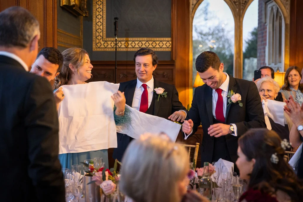 Wedding guests gathered around a banquet table in a grand manor house laugh joyfully during the speeches.