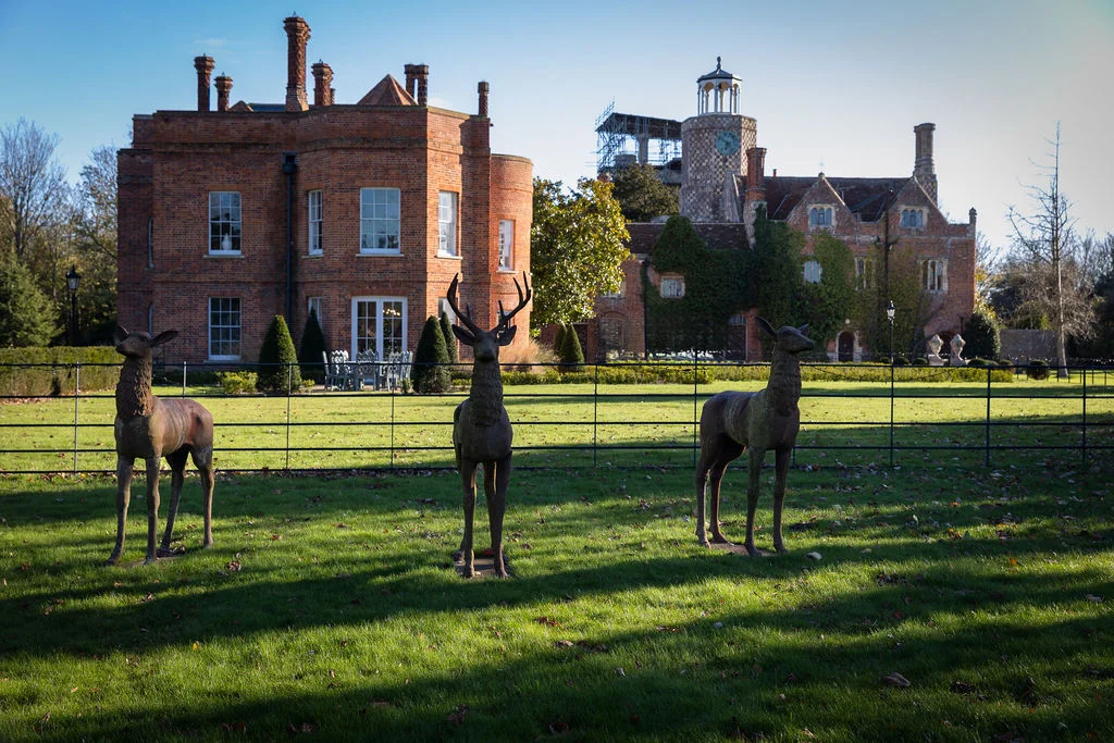 An exterior view of a historic red brick country house wedding venue featuring a manicured lawn and three decorative metal deer statues in the foreground.