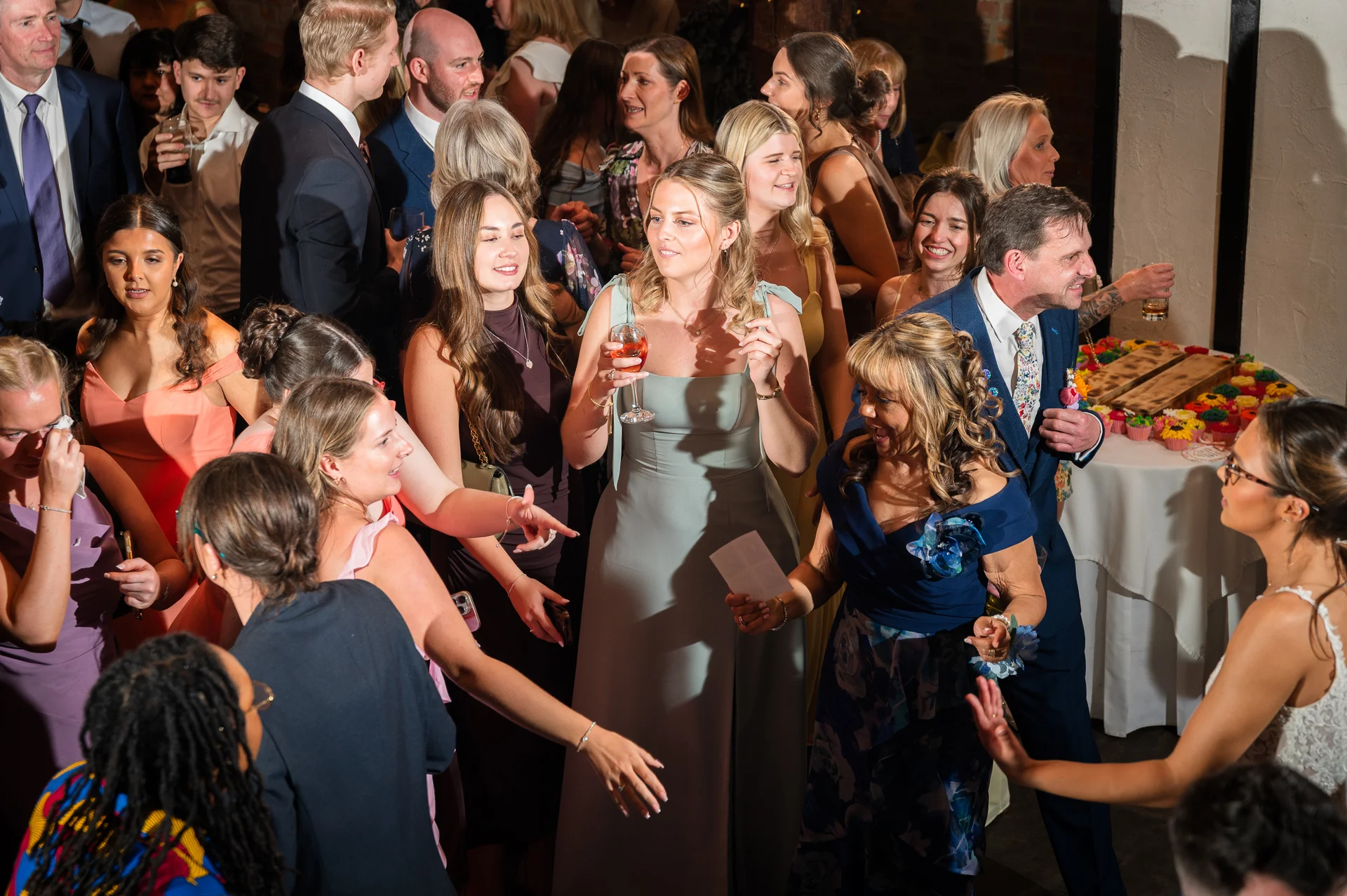 A lively group of wedding guests mingle and socialise indoors during the evening reception, holding drinks and chatting warmly under direct flash lighting.