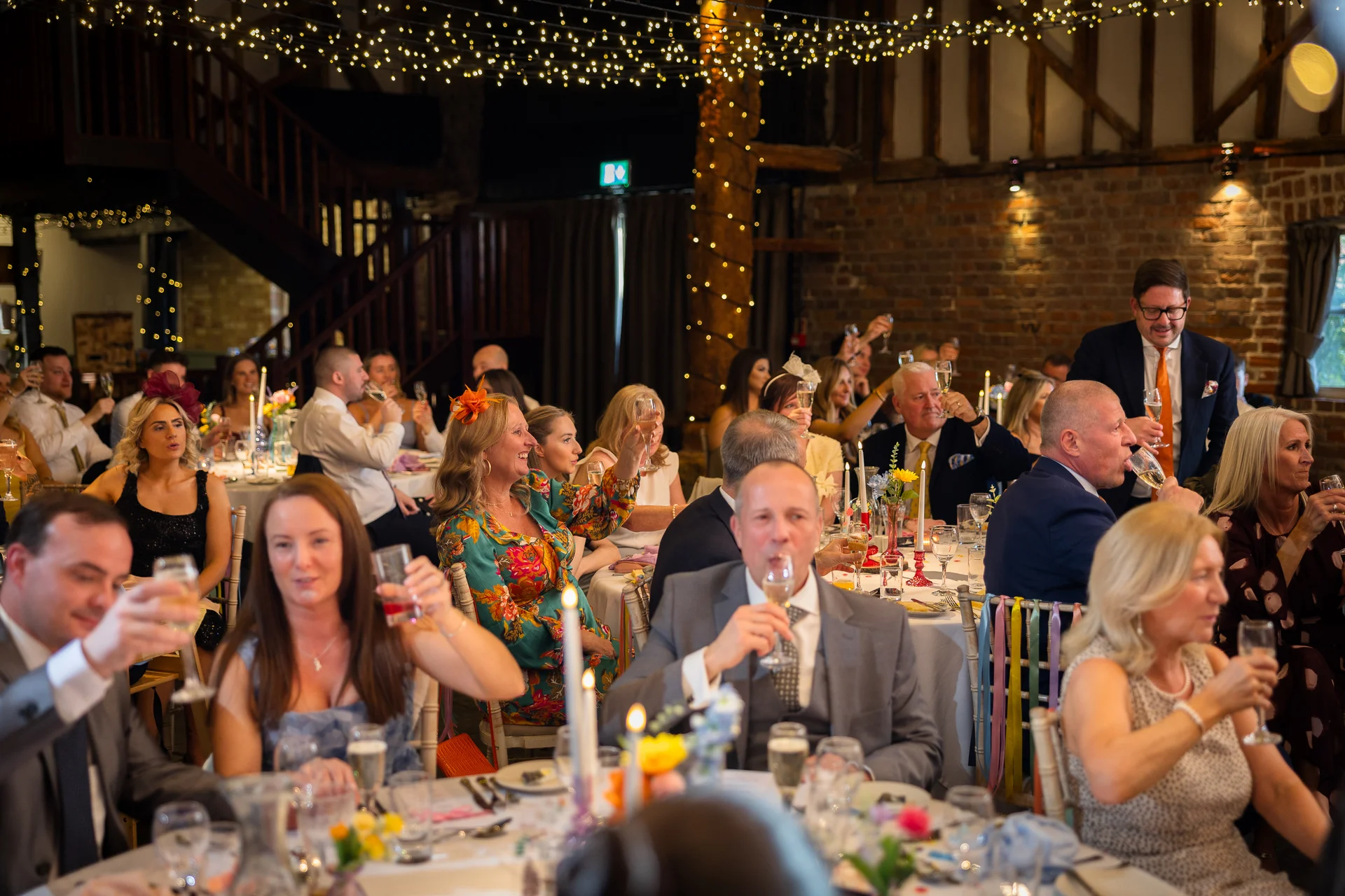 Wedding guests raise their glasses in a toast during the wedding breakfast inside a rustic barn venue decorated with exposed brick and fairy lights.
