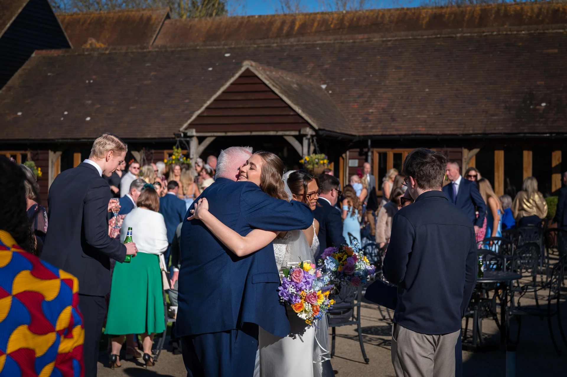A bride embraces her father warmly in a sunlit courtyard outside a rustic barn while wedding guests mingle in the background.