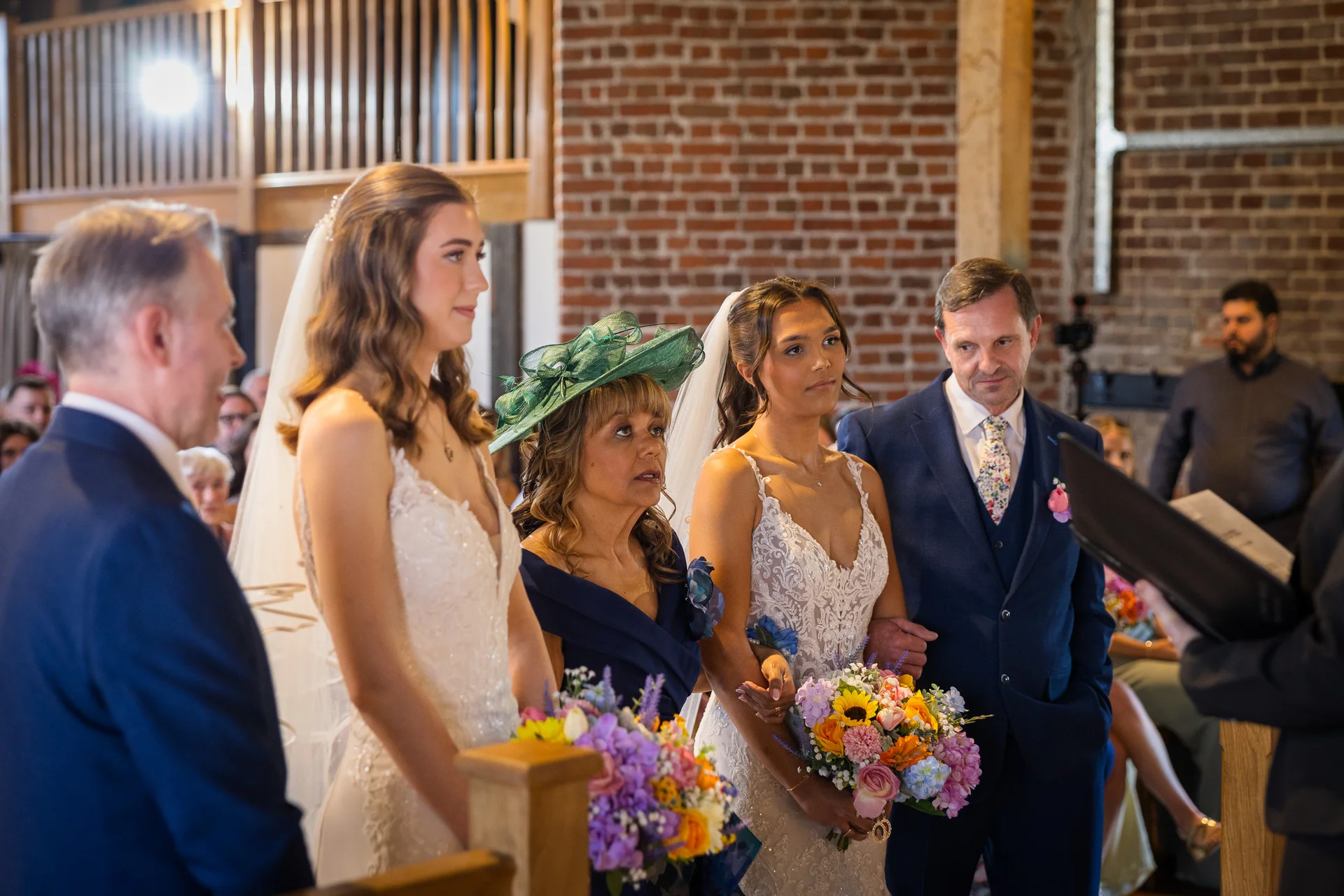 A bride, bridesmaids in matching lace gowns, and family members stand together inside a rustic brick barn during a wedding ceremony.