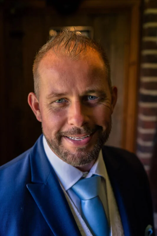 A close-up portrait of a groom wearing a blue suit and matching blue tie, smiling warmly toward the camera.