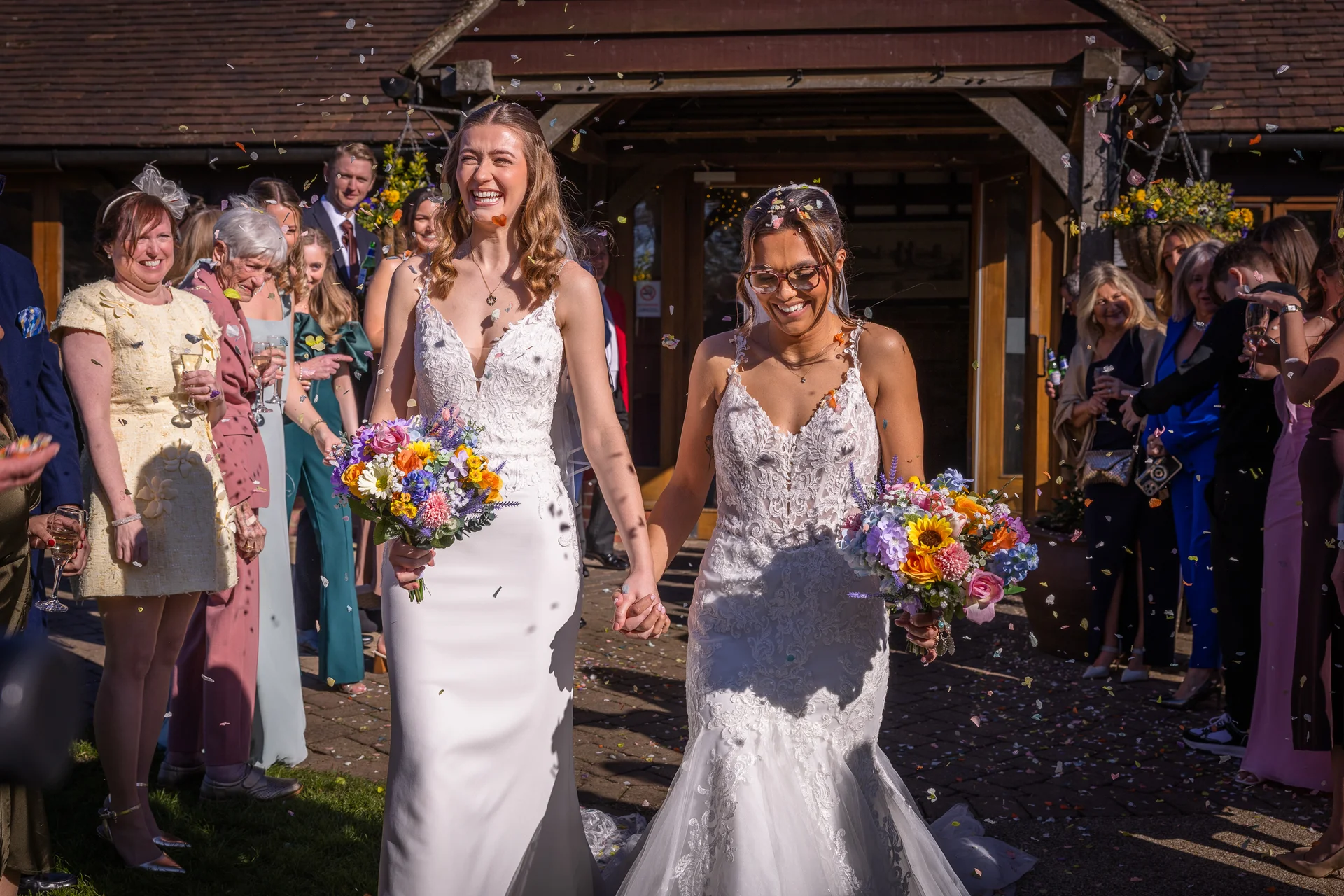 Two smiling brides holding hands and carrying colourful wildflower bouquets walk out of a stone barn venue while guests shower them with confetti in the bright sunshine.