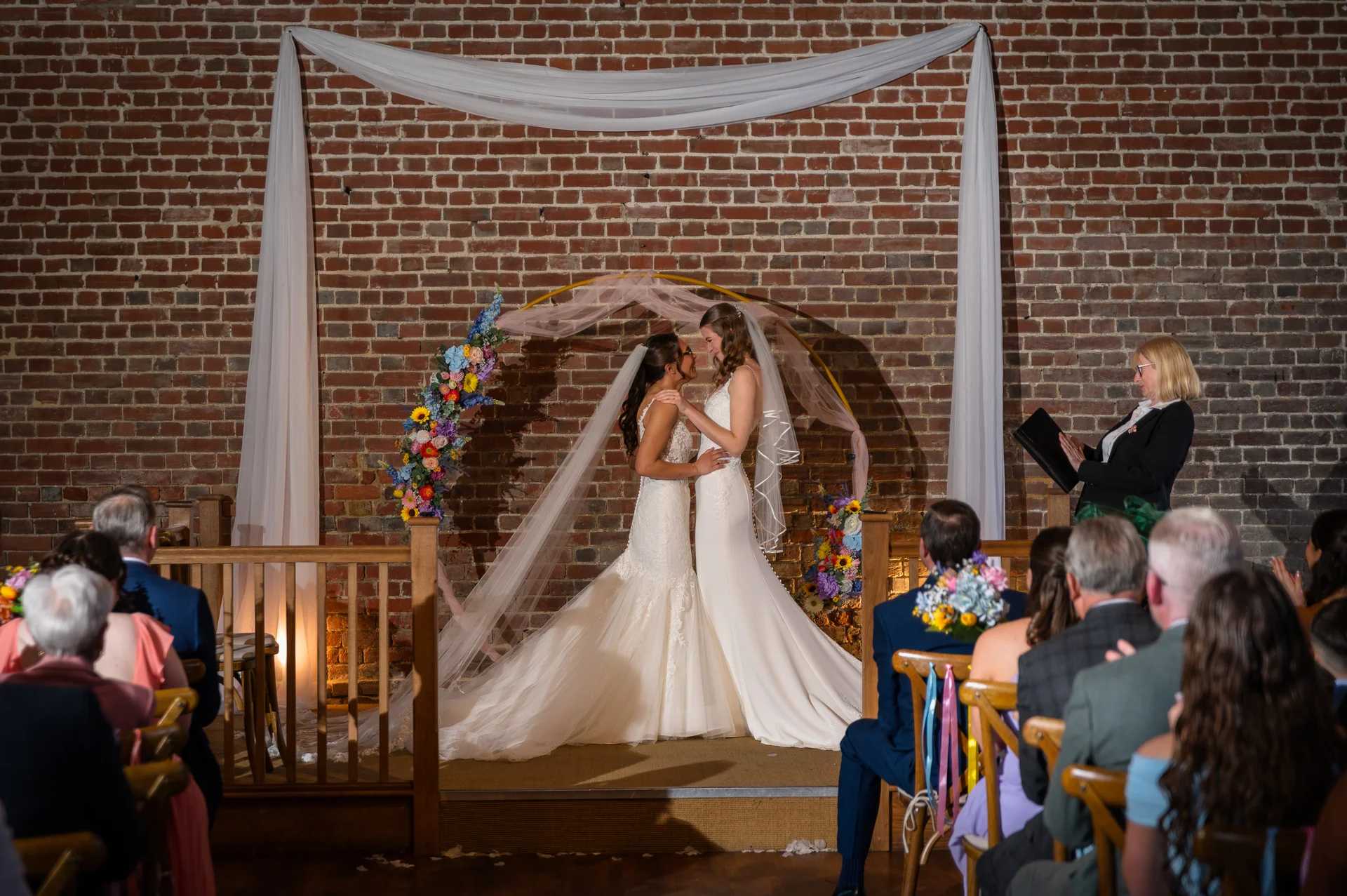 Two brides in white wedding dresses share a tender moment during an indoor ceremony framed by a floral arch and exposed brick wall.