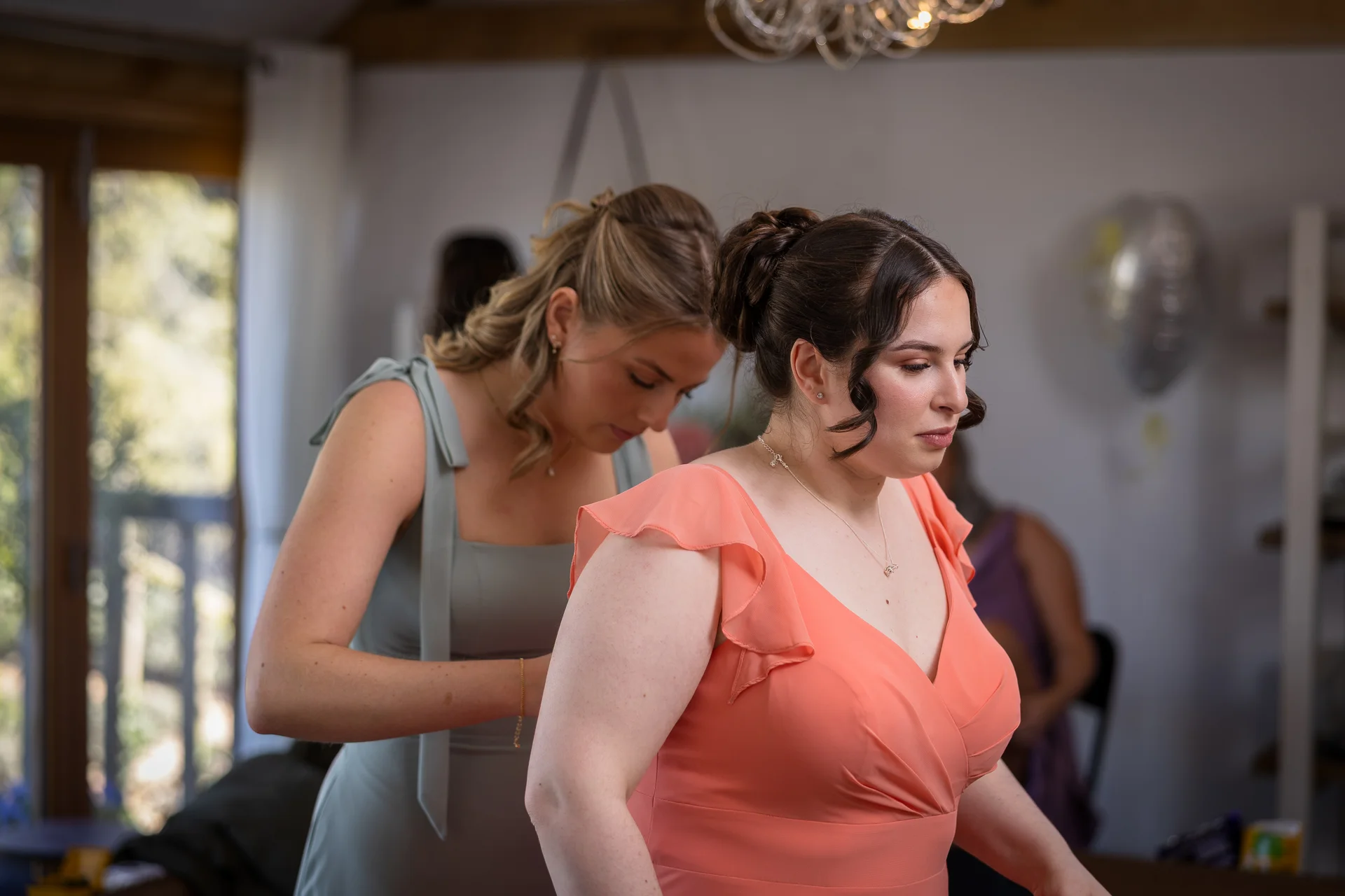 A bridesmaid in a sage green dress helps another woman in a peach dress with finishing touches during wedding morning preparations.