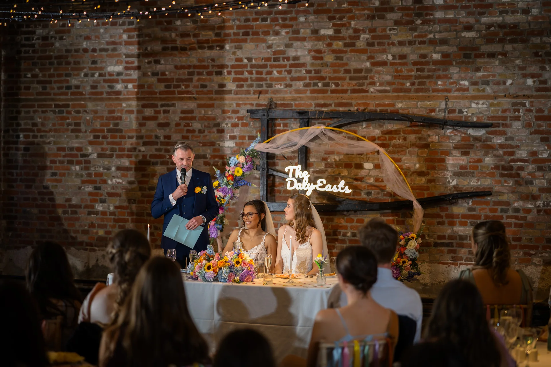 A guest delivers a speech with a microphone at the top table while two brides listen attentively in front of a rustic exposed brick wall and neon sign.