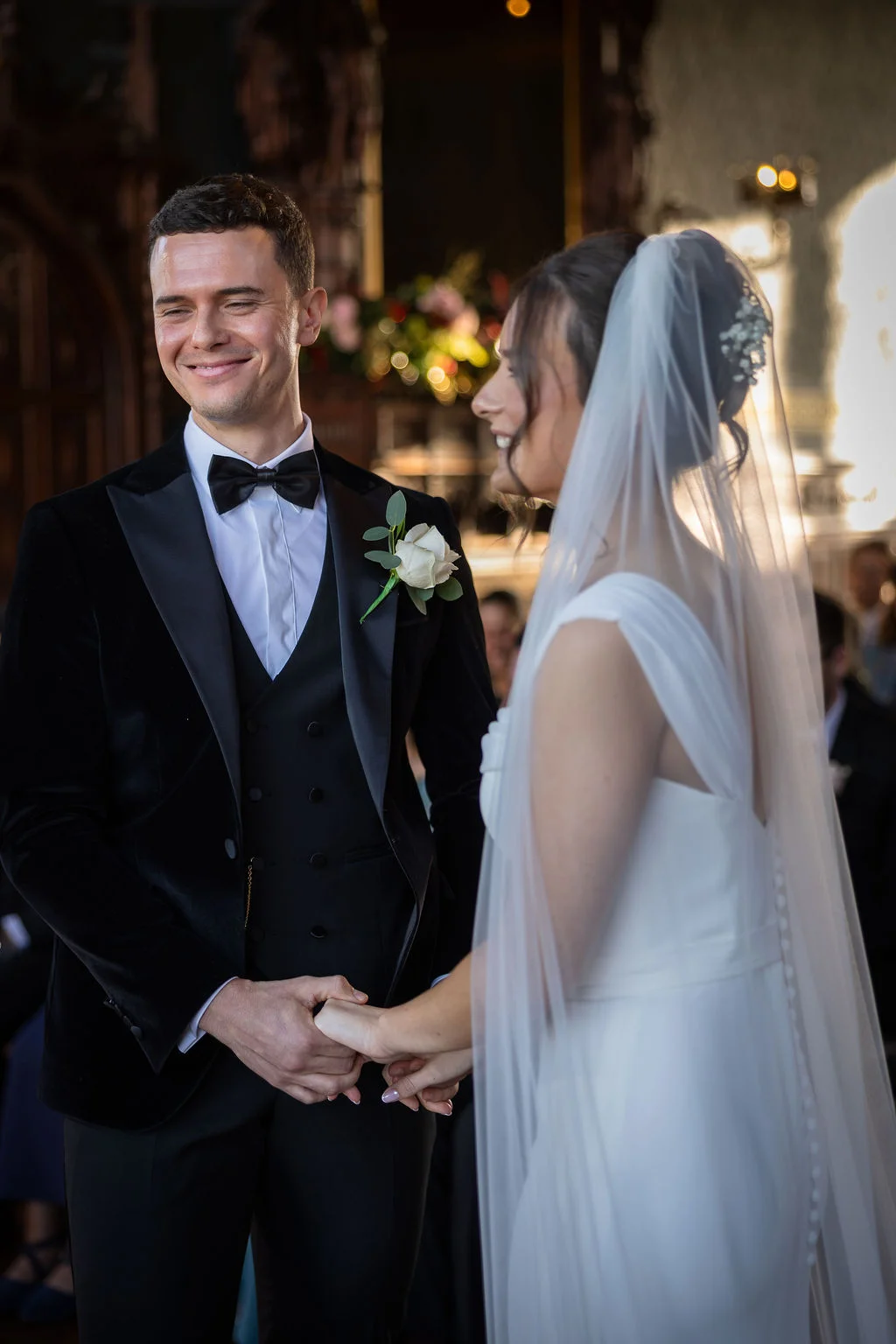 A smiling groom in a black velvet tuxedo and a bride in a white gown hold hands during their wedding ceremony.
