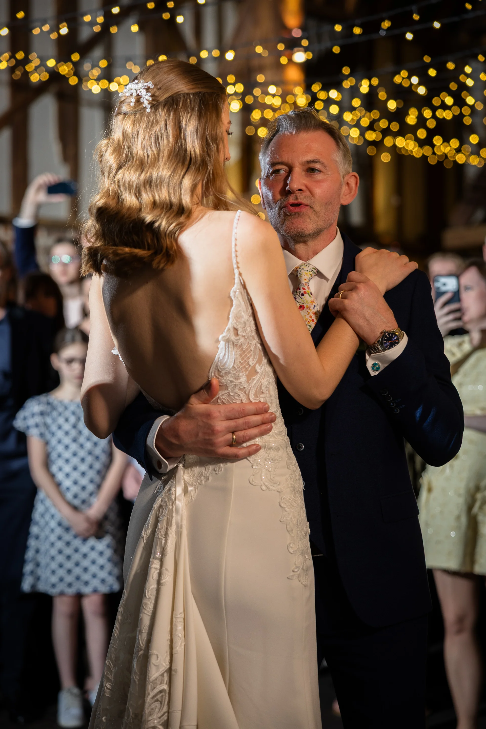 A father and bride share an emotional dance together beneath a glowing canopy of fairy lights inside a rustic timber barn.