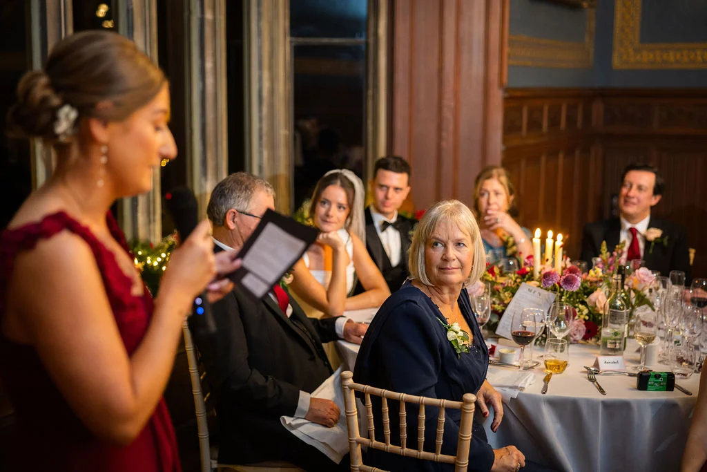 A bridesmaid delivers a speech at a wedding reception while seated guests listen attentively around a candlelit table inside a panelled hall.