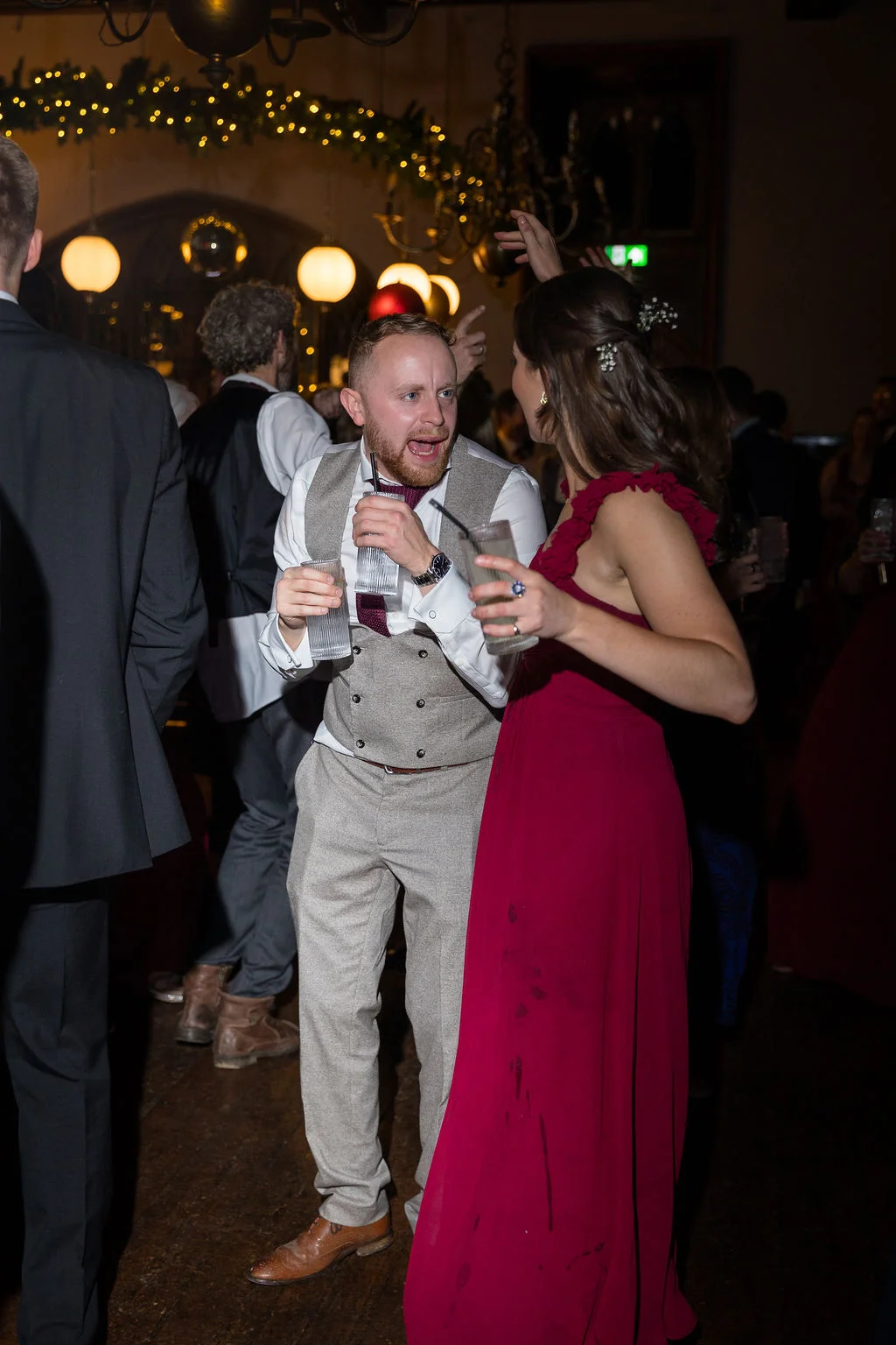 A groom in a tweed waistcoat and a guest in a burgundy dress enjoy a lively moment with drinks on a crowded wedding dance floor.