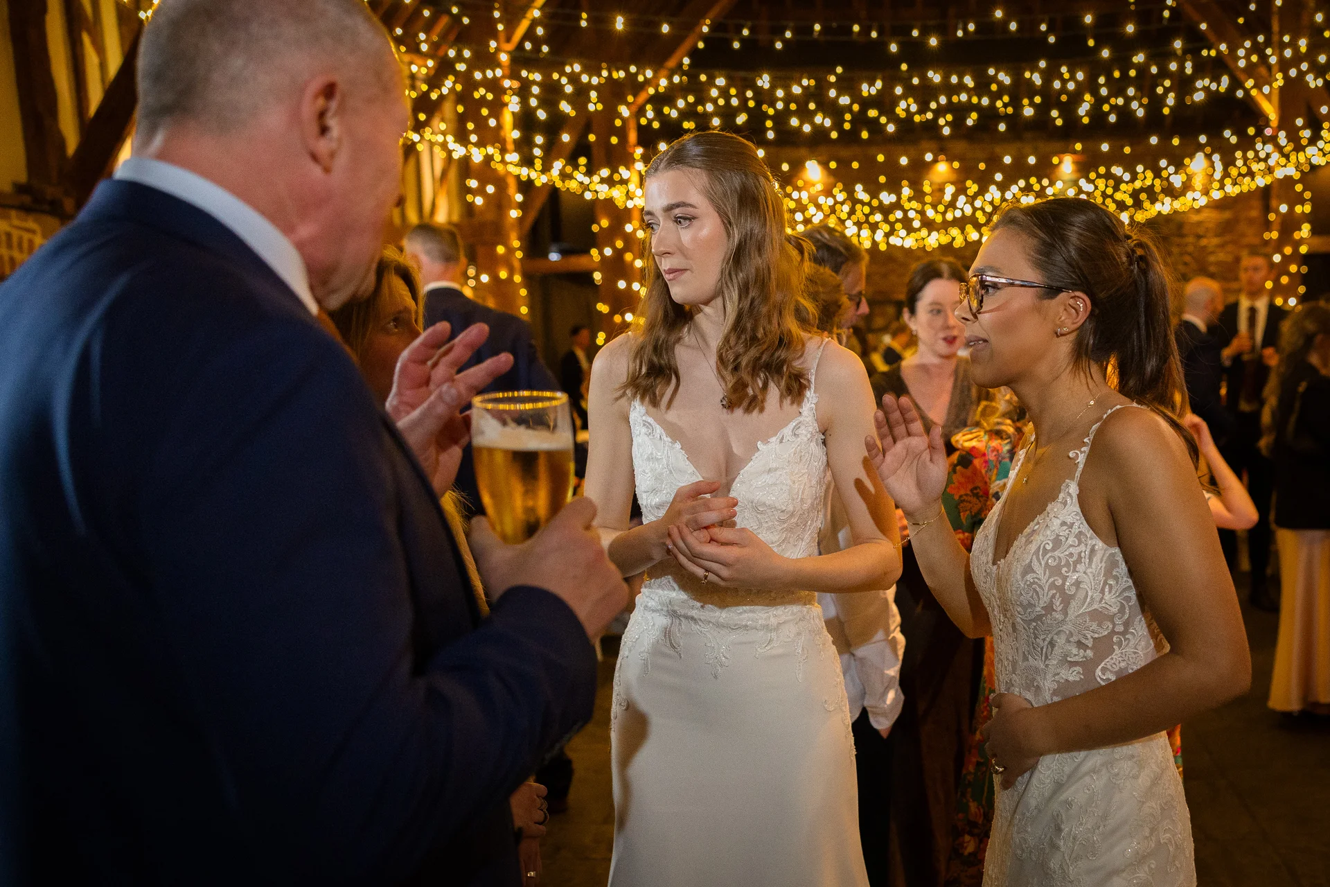 Two brides in lace-detailed wedding dresses chat animatedly with wedding guests inside a rustic barn illuminated by a warm canopy of fairy lights.
