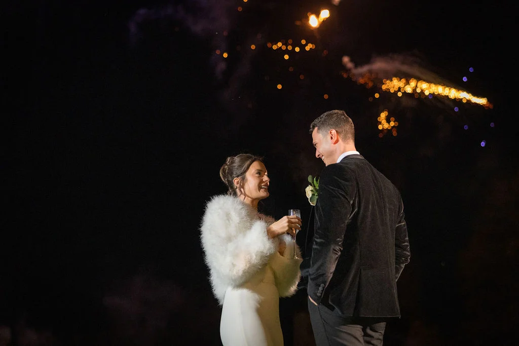 A newlywed couple stands outside at night, laughing together while holding champagne glasses beneath a dazzling display of fireworks.