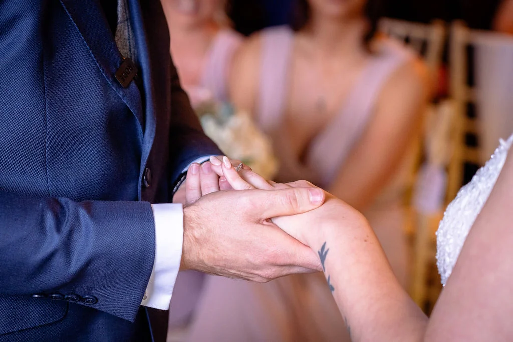 A groom in a navy suit places a wedding ring onto the hand of a bride wearing a lace dress during their ceremony.