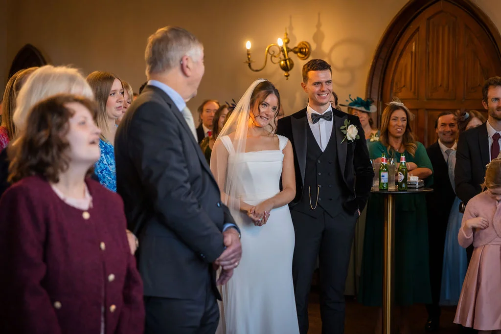 A smiling bride in a modern white dress with a veil stands beside her groom in a classic black tuxedo during an indoor wedding ceremony.