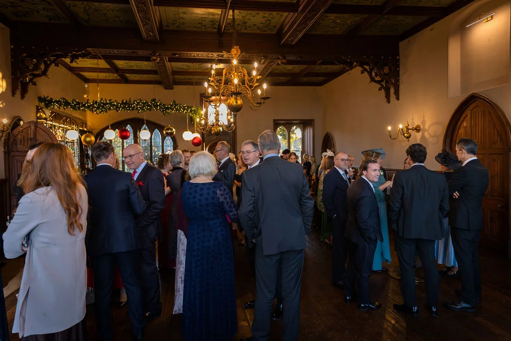 Wedding guests mingle during a drinks reception inside a historic wood-panelled manor house reception room illuminated by a grand chandelier.