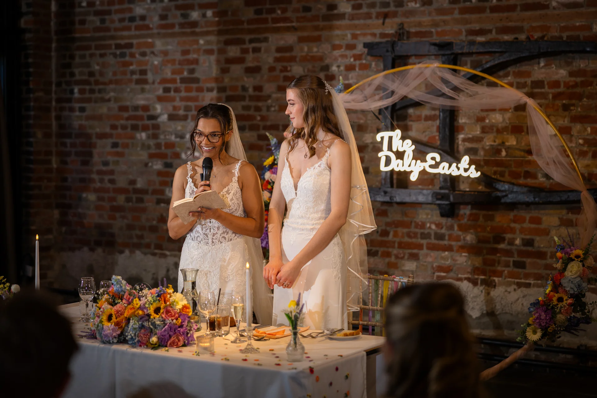 One bride gives a speech holding a microphone and a notebook at the top table while her smiling partner stands beside her in front of an exposed brick wall and a custom neon sign.