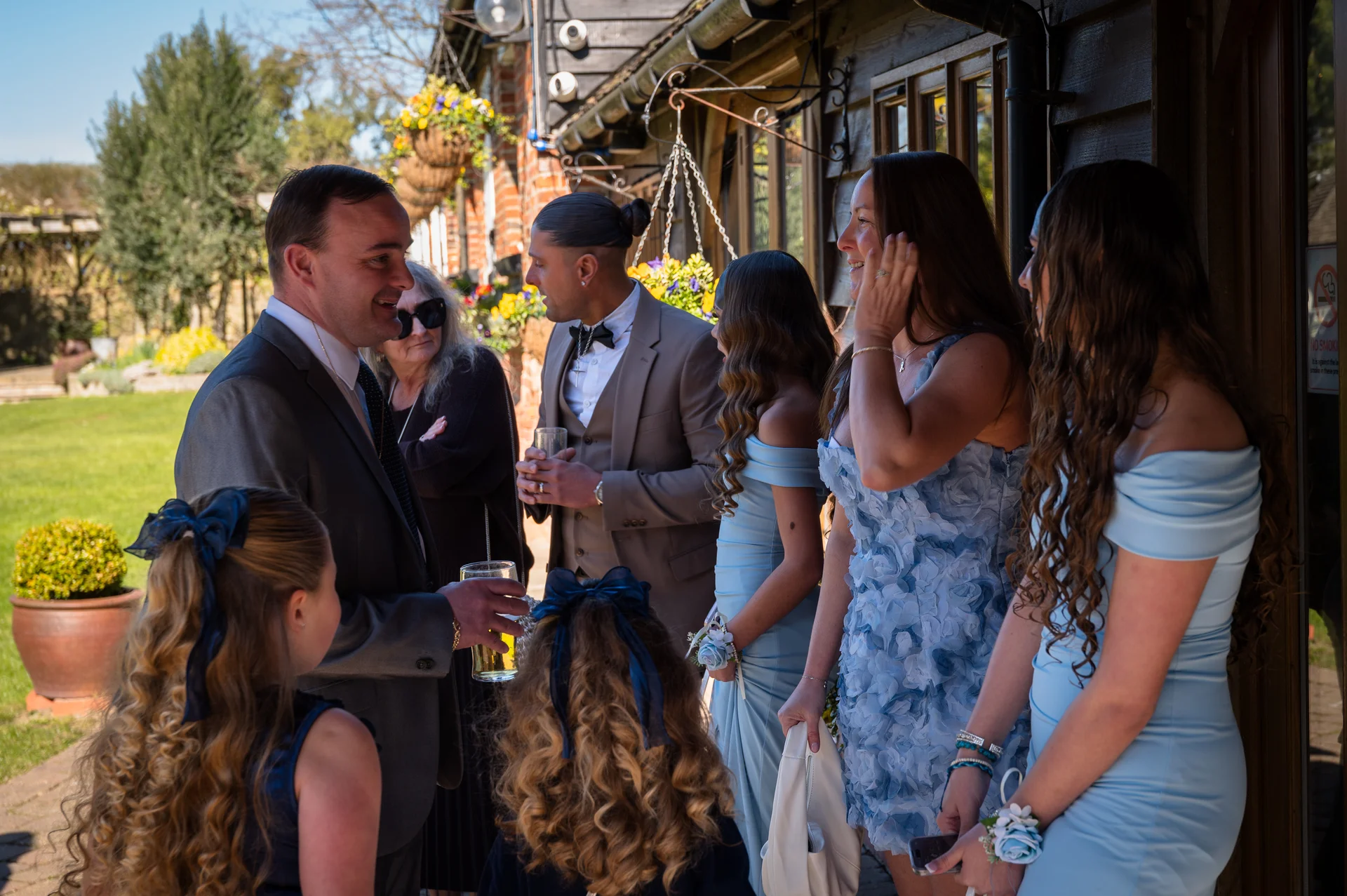 Wedding guests and bridesmaids in blue dresses mingle outdoors in the sunshine near a rustic building.