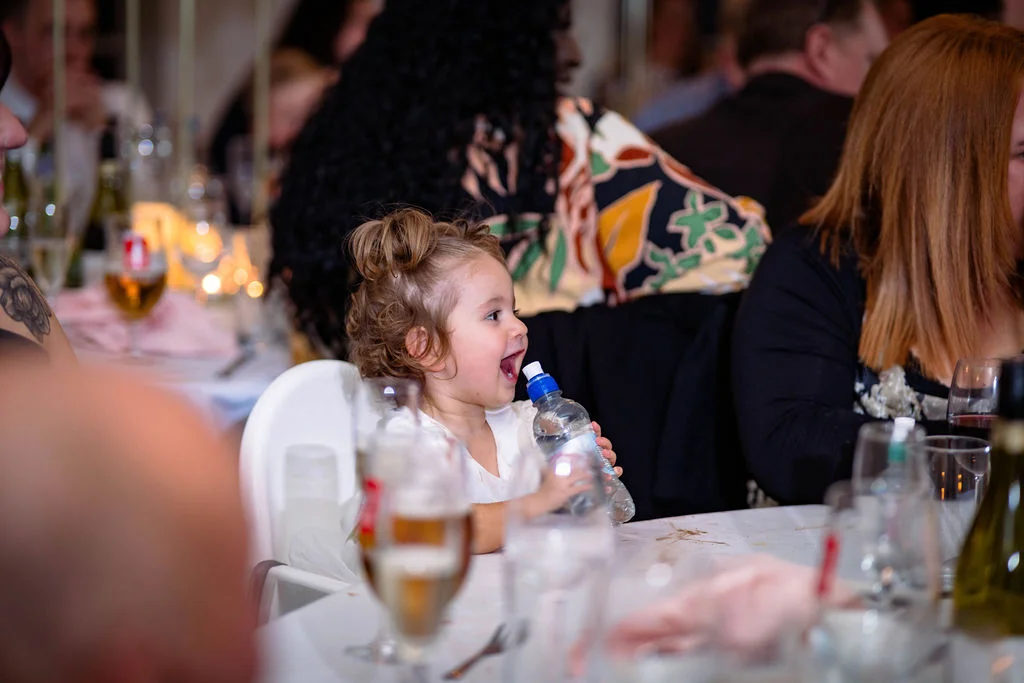 A smiling young child holding a water bottle at a wedding reception table surrounded by guests.