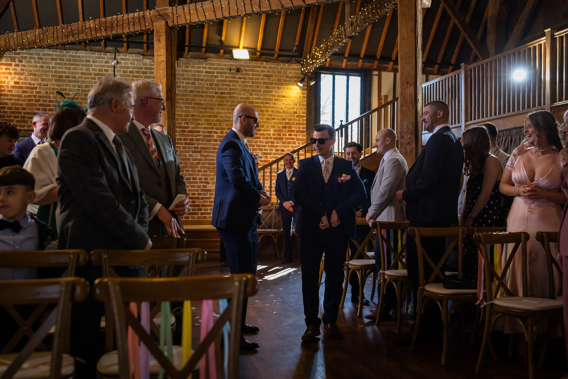 The groom and his groomsmen wear matching navy three-piece suits while waiting at the altar inside a rustic barn venue decorated with wooden beams and fairy lights.