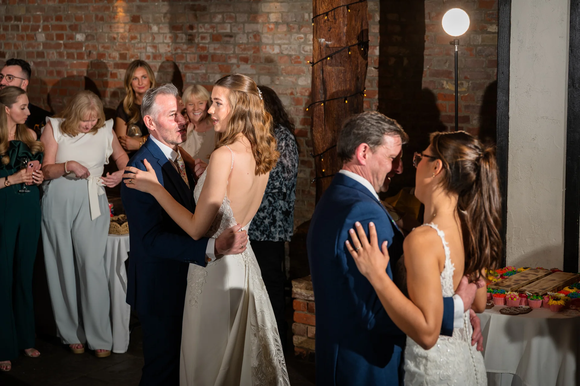 Couples share a joyful first dance inside a rustic barn venue with exposed brick walls and warm lighting.