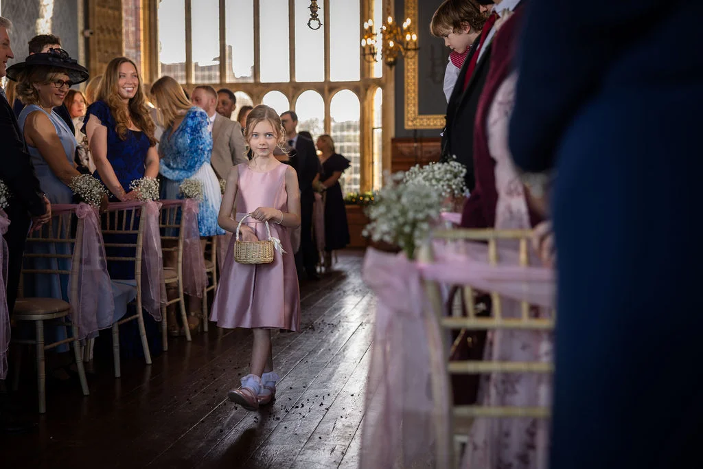 A young flower girl in a pink dress walks down the aisle carrying a small basket inside a historic church with large windows.
