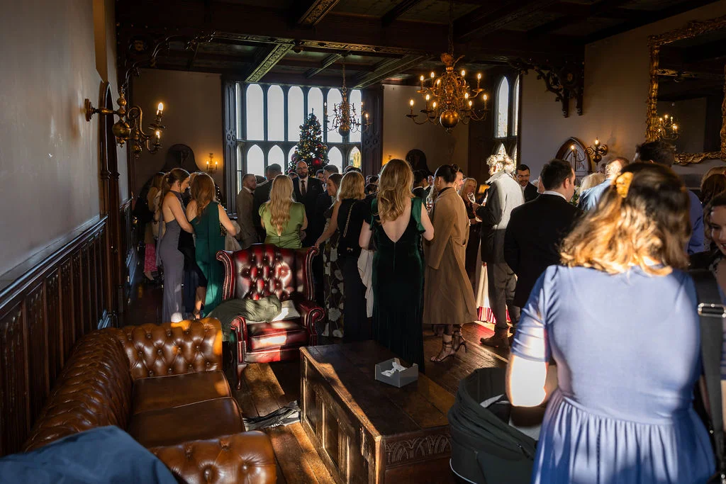 Wedding guests mingle during a festive drinks reception inside a grand, wood-panelled manor house room illuminated by warm afternoon sunlight.