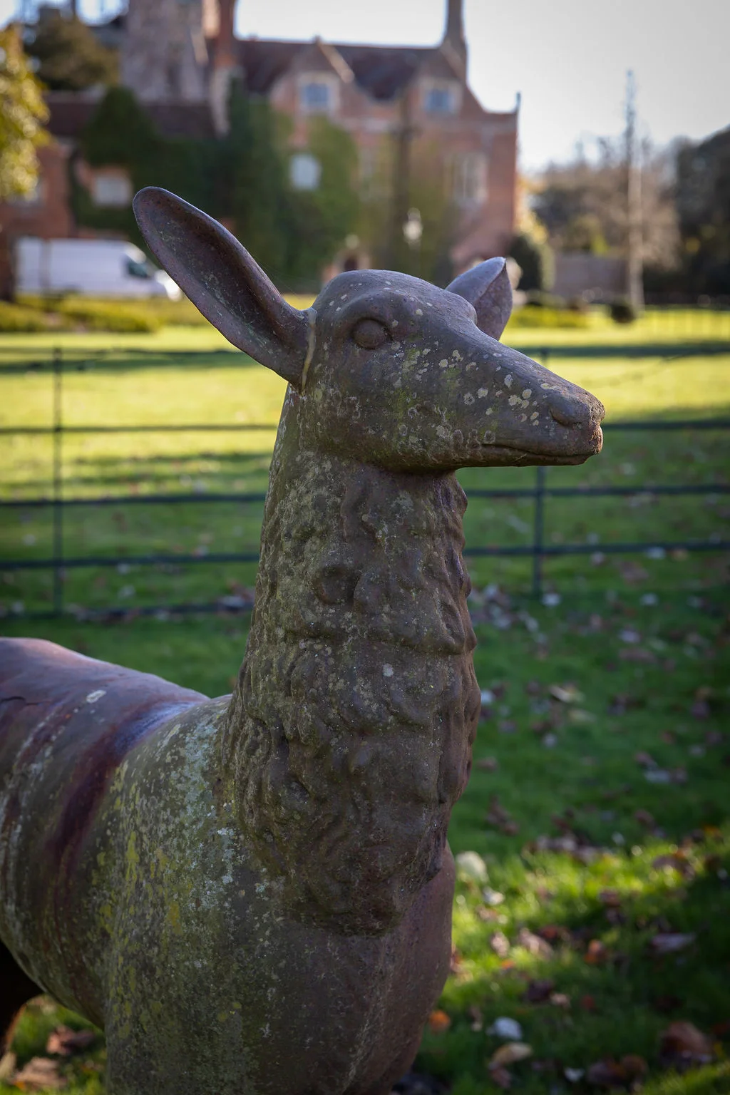 A weathered metal deer sculpture stands on the manicured lawn of a grand historic country house venue on a bright afternoon.