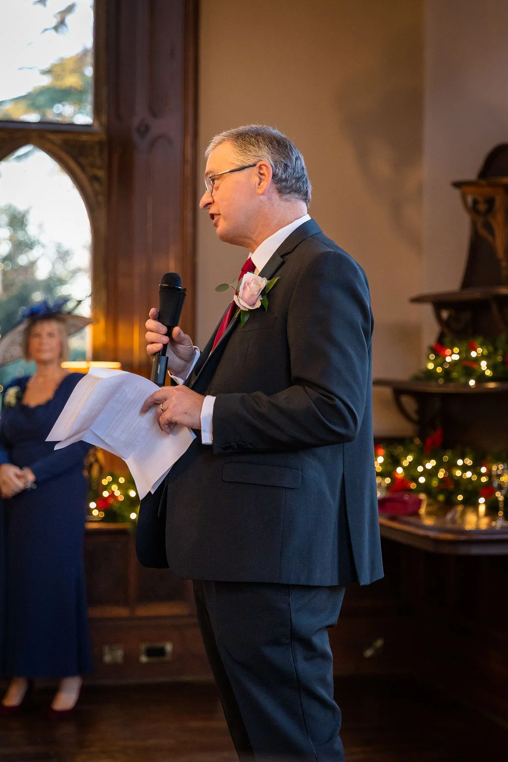 A groom wearing a charcoal suit delivers his wedding speech, holding a microphone and notes with festive fairy lights glowing in the background.