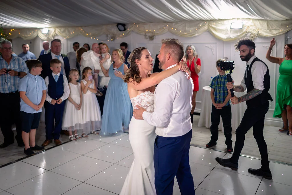 Newlyweds share their first dance on a lit white dance floor inside a marquee while guests and a videographer watch.