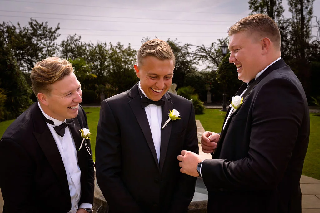 Three groomsmen in classic black tuxedos and bow ties share a joyful laughing moment outdoors on a sunny wedding day.