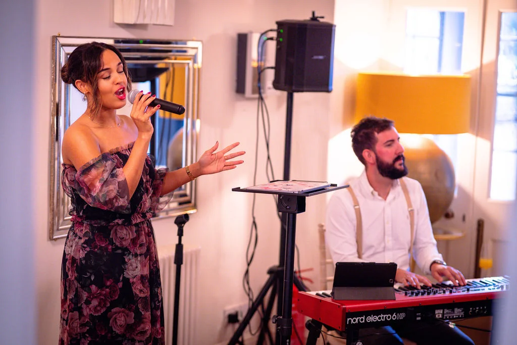 A female vocalist in a floral print off-the-shoulder dress sings into a microphone while a pianist in braces plays a red keyboard at an indoor wedding reception.