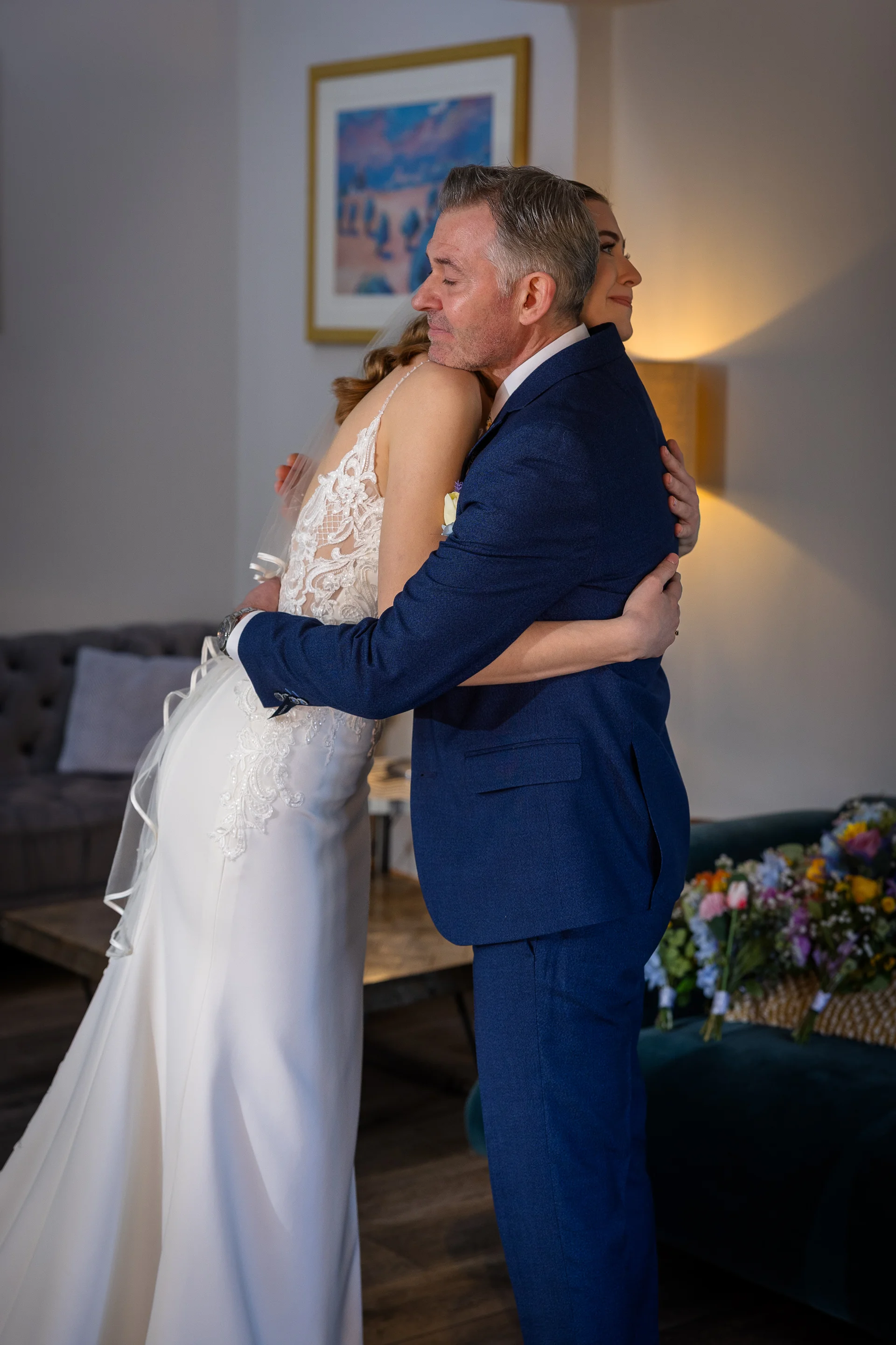 An emotional father wearing a navy blue suit shares a warm, tender hug with his daughter who is dressed in a white lace wedding gown and veil.