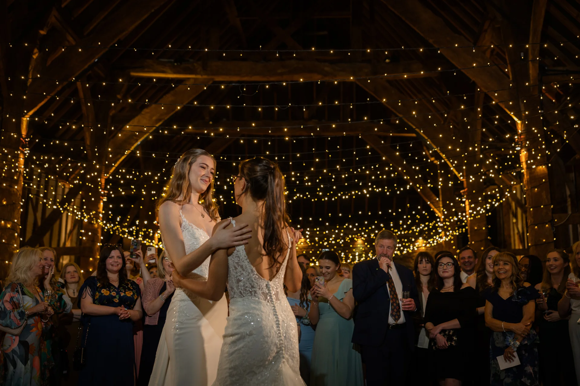 Two brides share their first dance beneath a breathtaking canopy of fairy lights inside a rustic oak-framed barn surrounded by smiling guests.