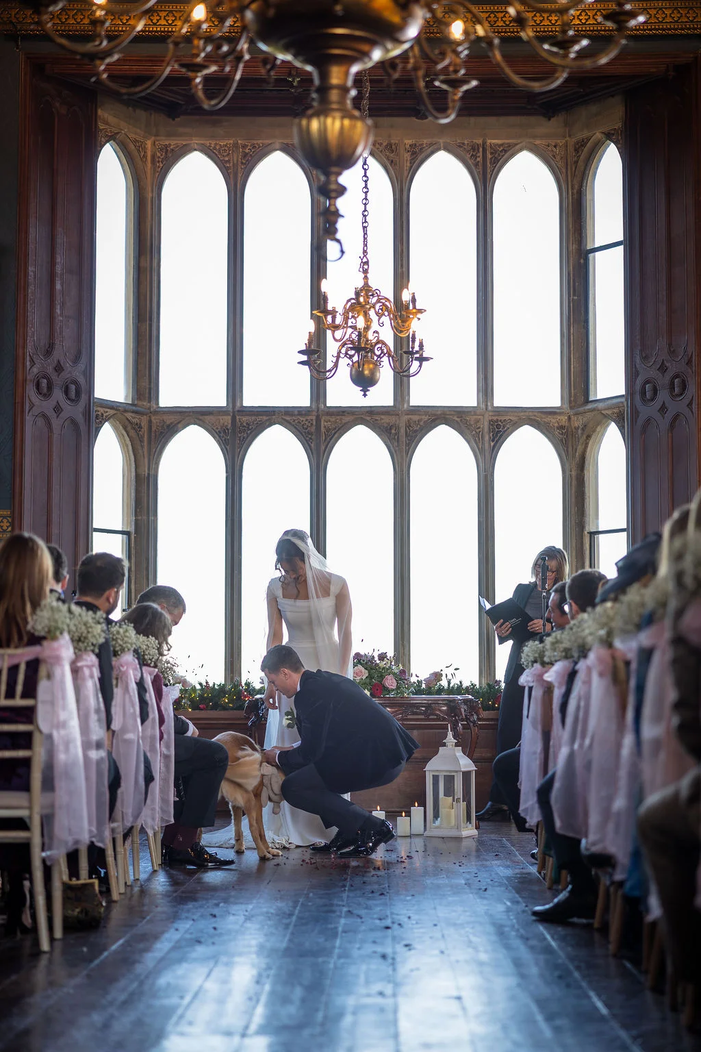 A groom kneels to pet a dog at the front of a grand ceremony room featuring towering stained-glass windows and elegant wooden flooring.
