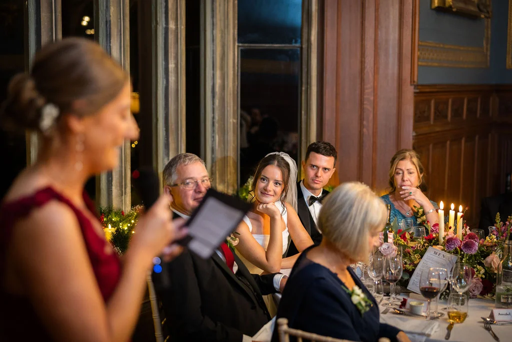 A guest delivers a speech at the top table during a candlelit wedding reception in a wood-panelled room, while the bride and groom listen attentively.
