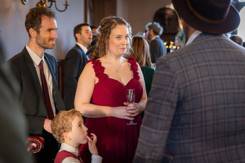 Wedding guests mingling indoors during a reception, featuring a woman in a burgundy dress holding a champagne glass while talking to a man in a checked jacket.