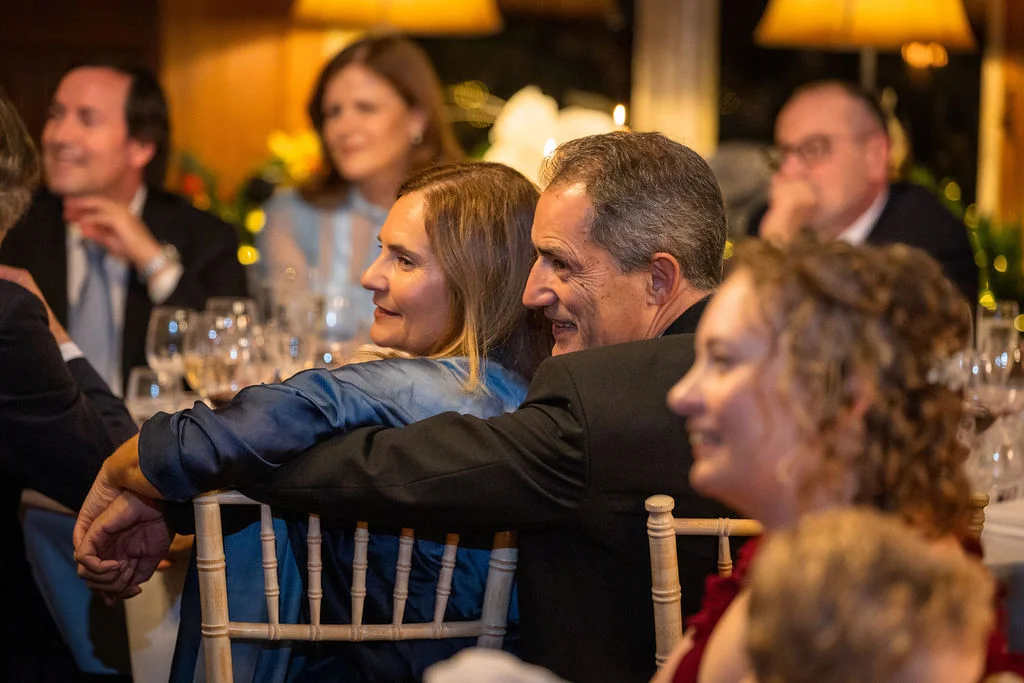An older couple shares a warm embrace while seated at a wedding reception table surrounded by smiling guests during the speeches.