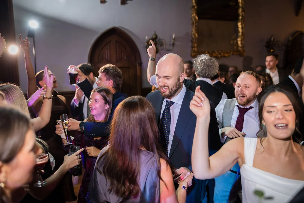A lively group of wedding guests smiling and dancing together on a crowded evening dance floor under warm indoor lighting.