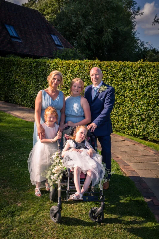 A happy wedding party, including adults in blue and a groom in a navy suit alongside children and a young girl in a wheelchair, posing outside in the sunshine.