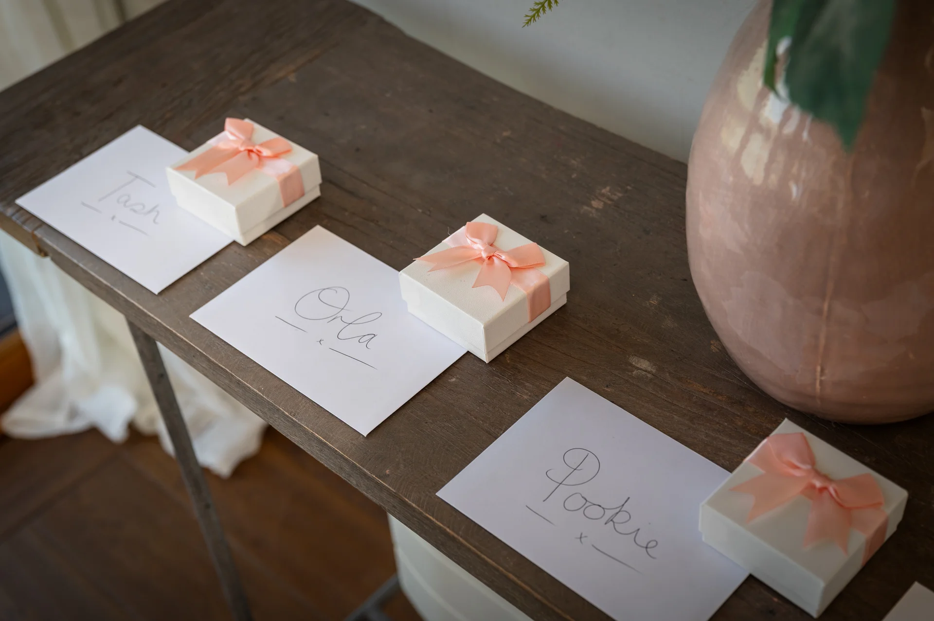 Personalised favour boxes with peach ribbons are neatly arranged alongside handwritten place cards on a rustic wooden console table.