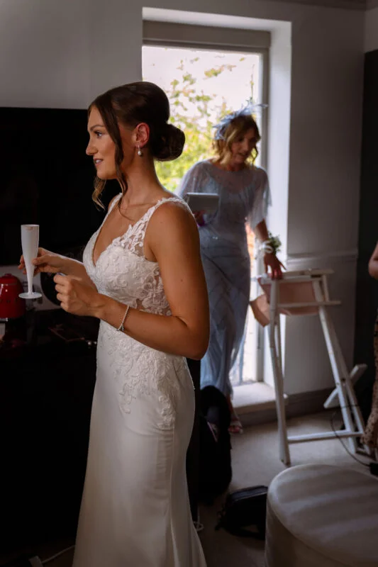 A bride in a fitted lace-detailed wedding gown holds a champagne flute indoors while a family member enters through a sunlit doorway behind her.