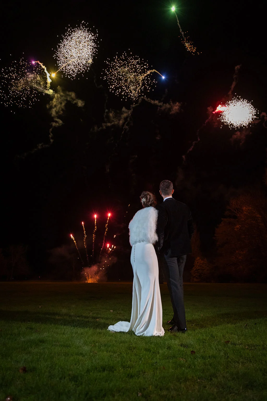 Newlyweds stand outdoors on the grass at night, watching a spectacular fireworks display light up the sky above them.