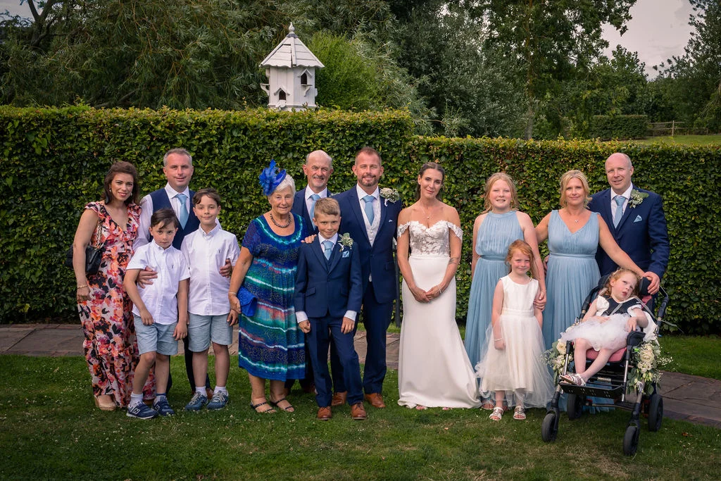 A large wedding party and family group pose together outdoors in a lush garden setting with a tall green hedge backdrop.