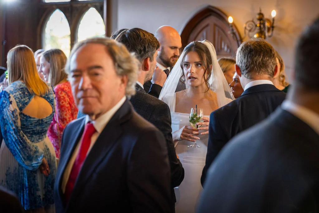 A candid shot captures the bride holding a garnished drink amidst a lively drinks reception, framed by guests mingling in a historic venue.