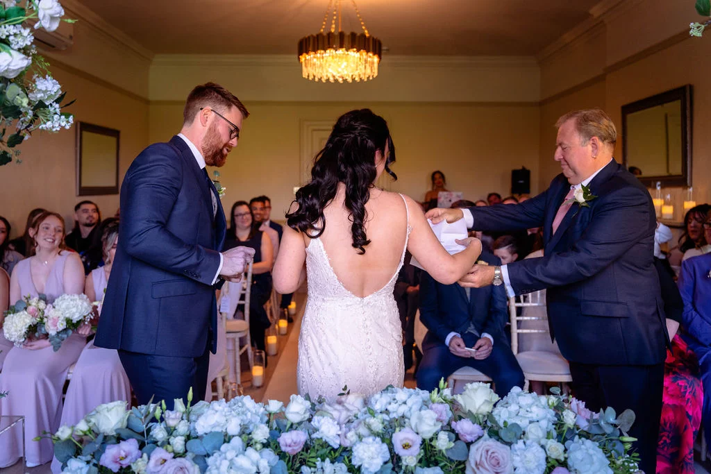 A bride and groom stand facing a registrar at the front of a light-filled ceremony room during the signing of the register, surrounded by floral arrangements and seated guests.